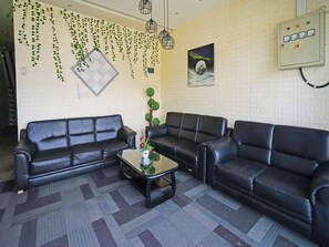 Sala de estar com sofás de couro preto, uma mesa de centro de vidro, plantas penduradas, um relógio de parede e uma tomada.