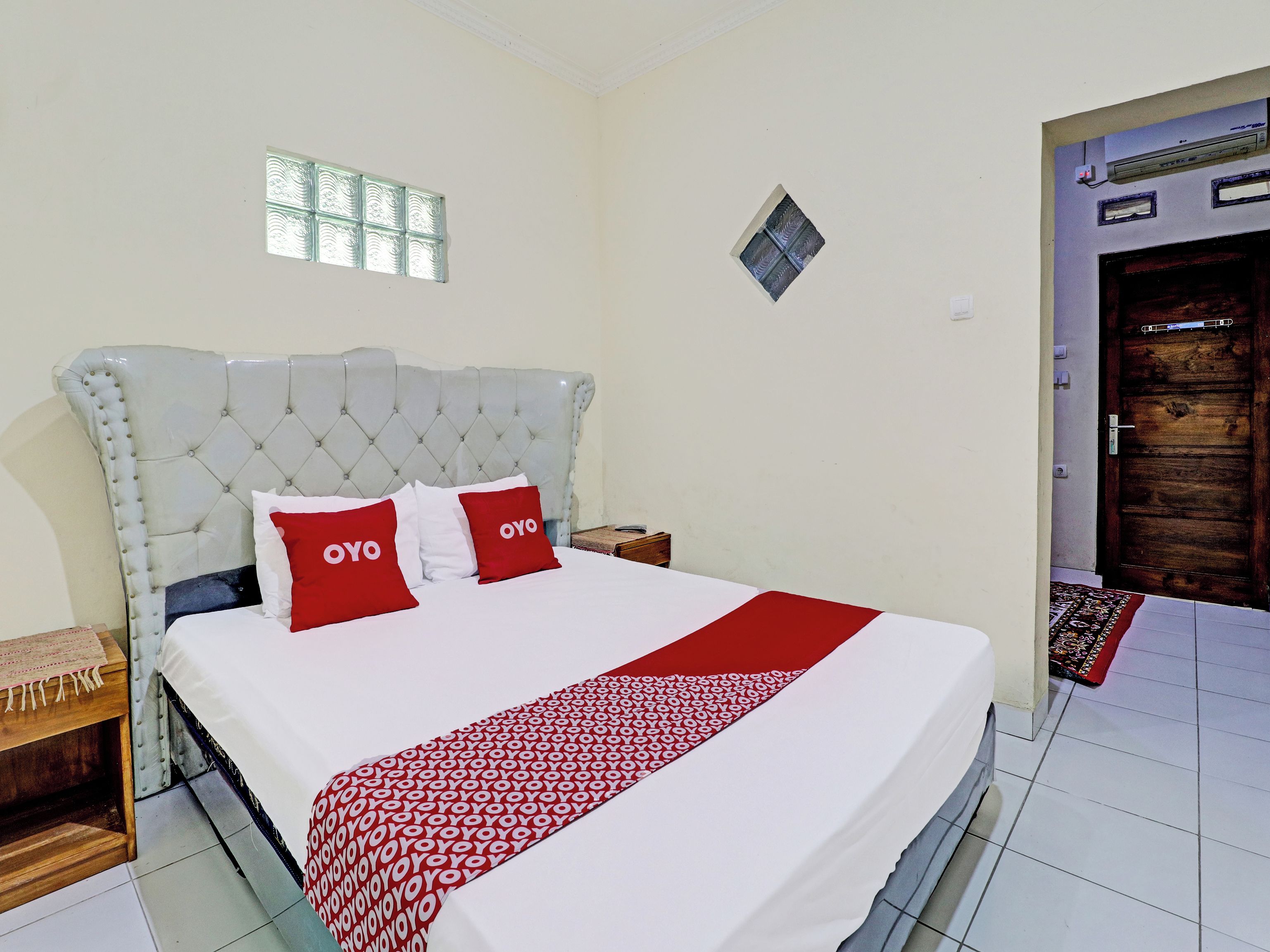 A bedroom with a tufted headboard, a bed with a white and red patterned bedspread, two red pillows with 'OYO' text, a small bedside table, and a window with a decorative glass panel.