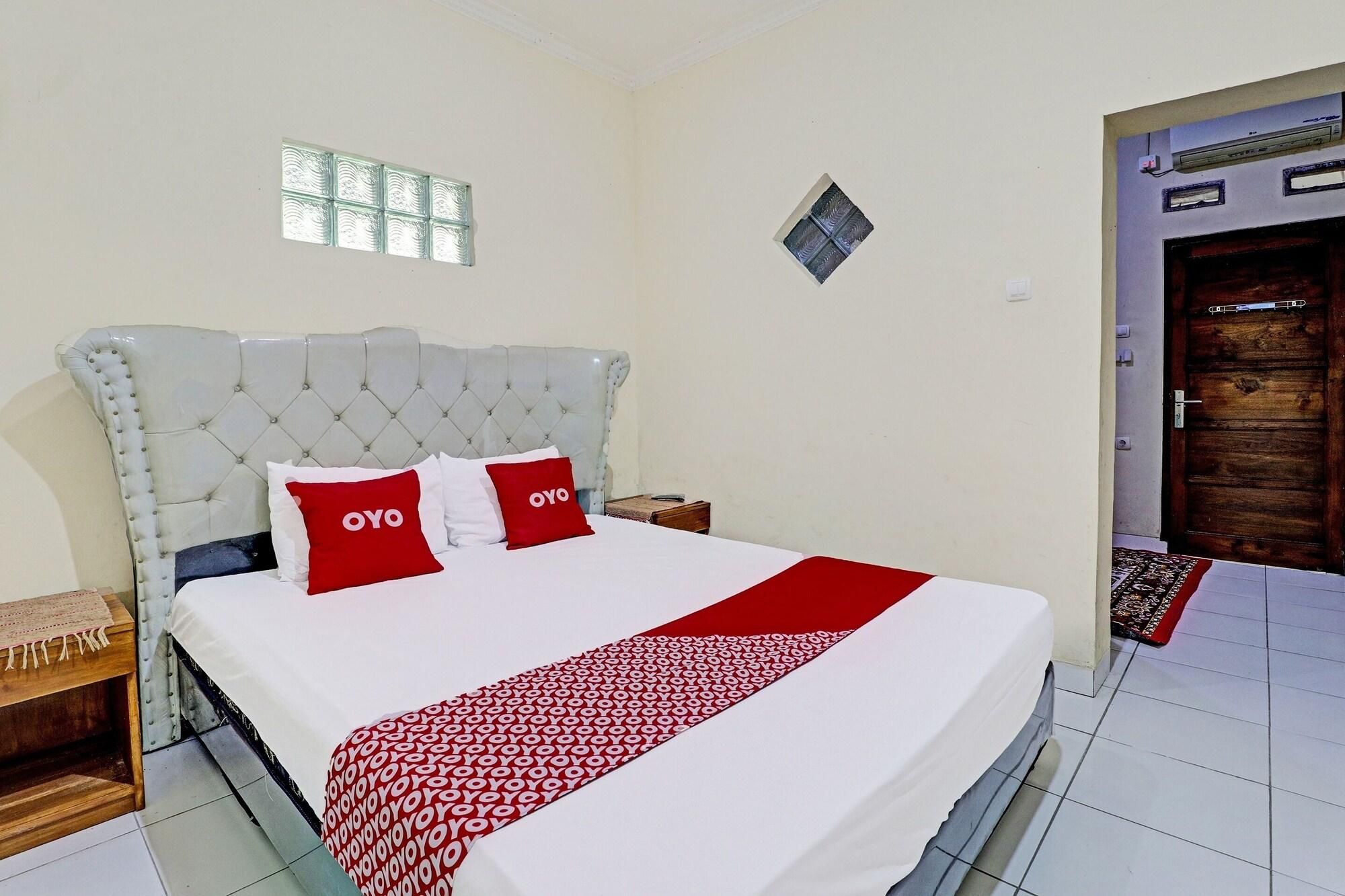 Habitación de hotel con una cama grande, dos almohadas rojas con la marca 'OYO', una colcha blanca y una manta estampada.