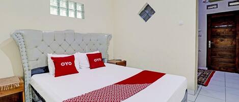 Habitación de hotel con una cama grande, dos almohadas rojas con la marca 'OYO', una colcha blanca y una manta estampada.