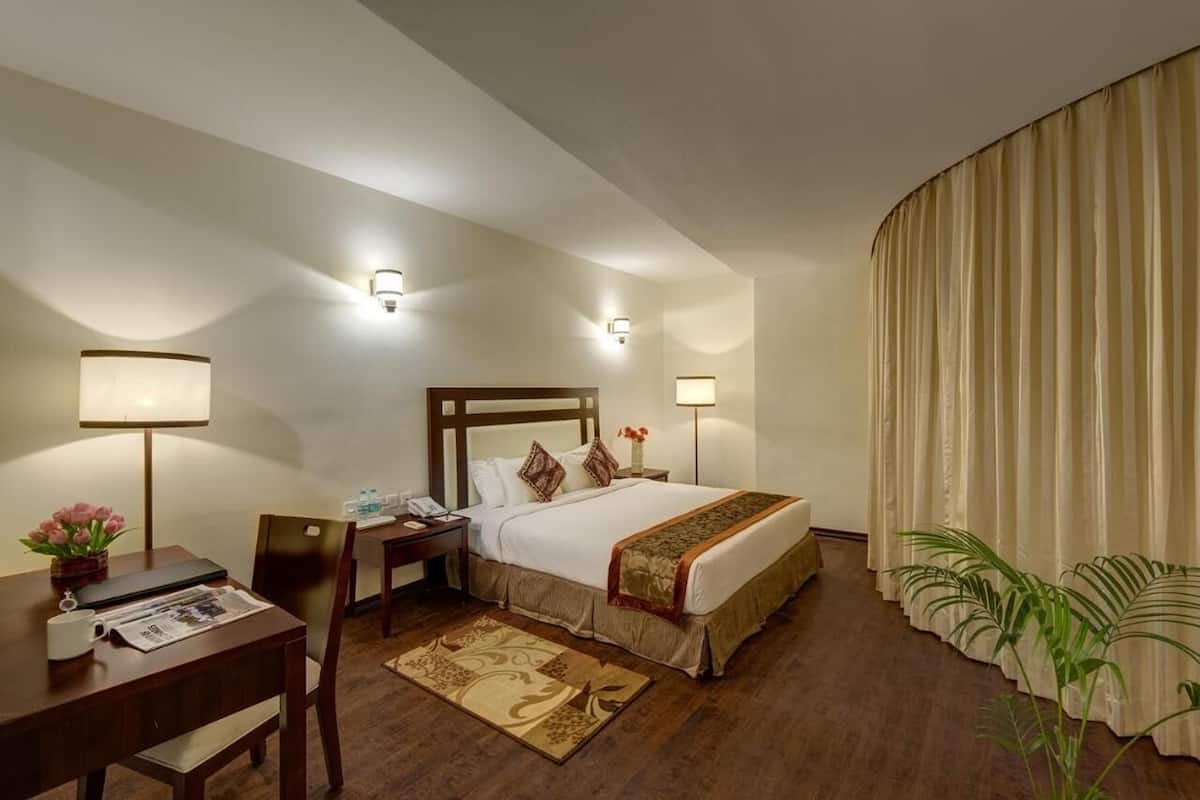 Habitación de hotel con cama, mesita de noche, lámpara, silla, escritorio y ventana con cortinas.