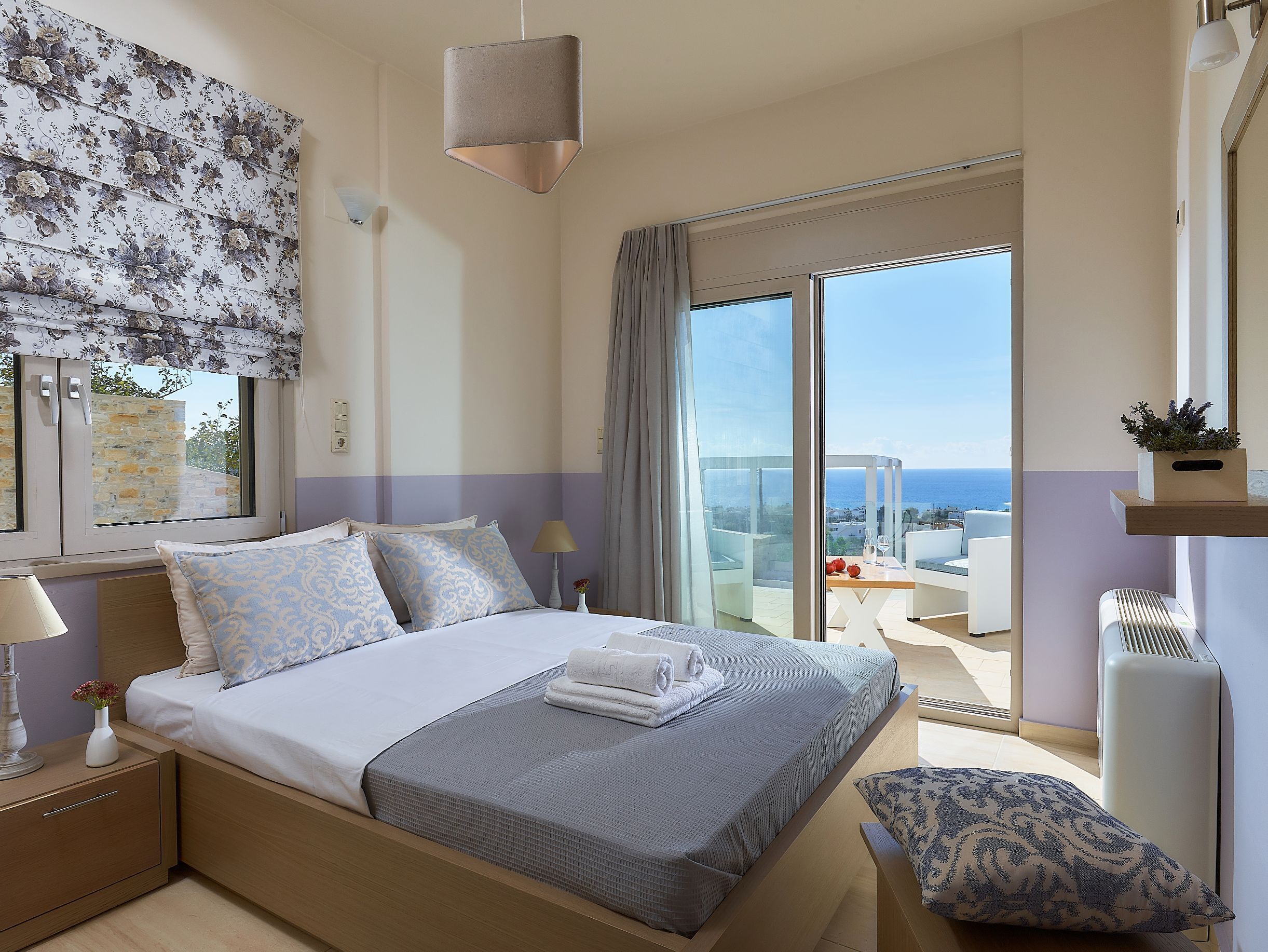 A modern hotel room with a bed, a nightstand, a lamp, a window with a view of the sea, and a balcony with a table and chairs.