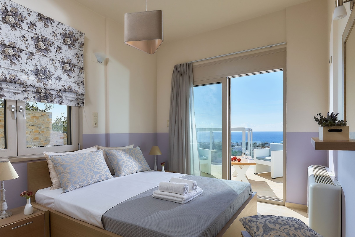 Ein modernes Hotelzimmer mit Bett, Nachttisch, Lampe, Fenster mit Meerblick und Balkon mit Tisch und Stühlen.