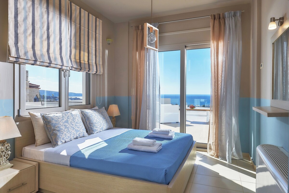 Ein Schlafzimmer mit Bett, Nachttischen, einer Lampe und Blick auf das Meer durch große Fenster.
