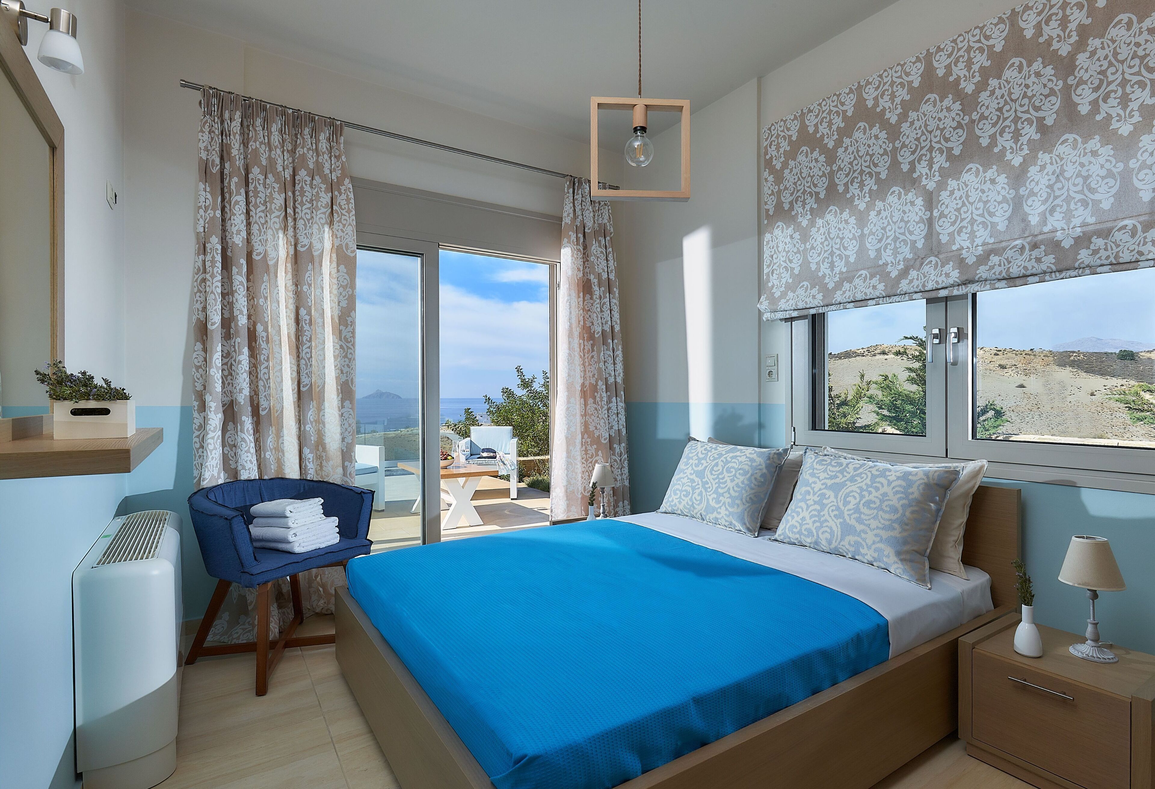 A bedroom with a large bed, a chair, a bedside table, a lamp, and a view of the outdoors.