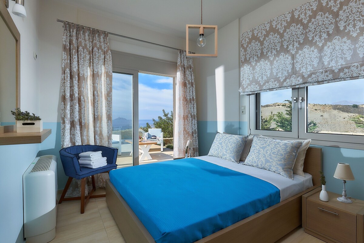 Ein Schlafzimmer mit einem großen Bett, einem Sessel, einem kleinen Tisch und Blick ins Freie.