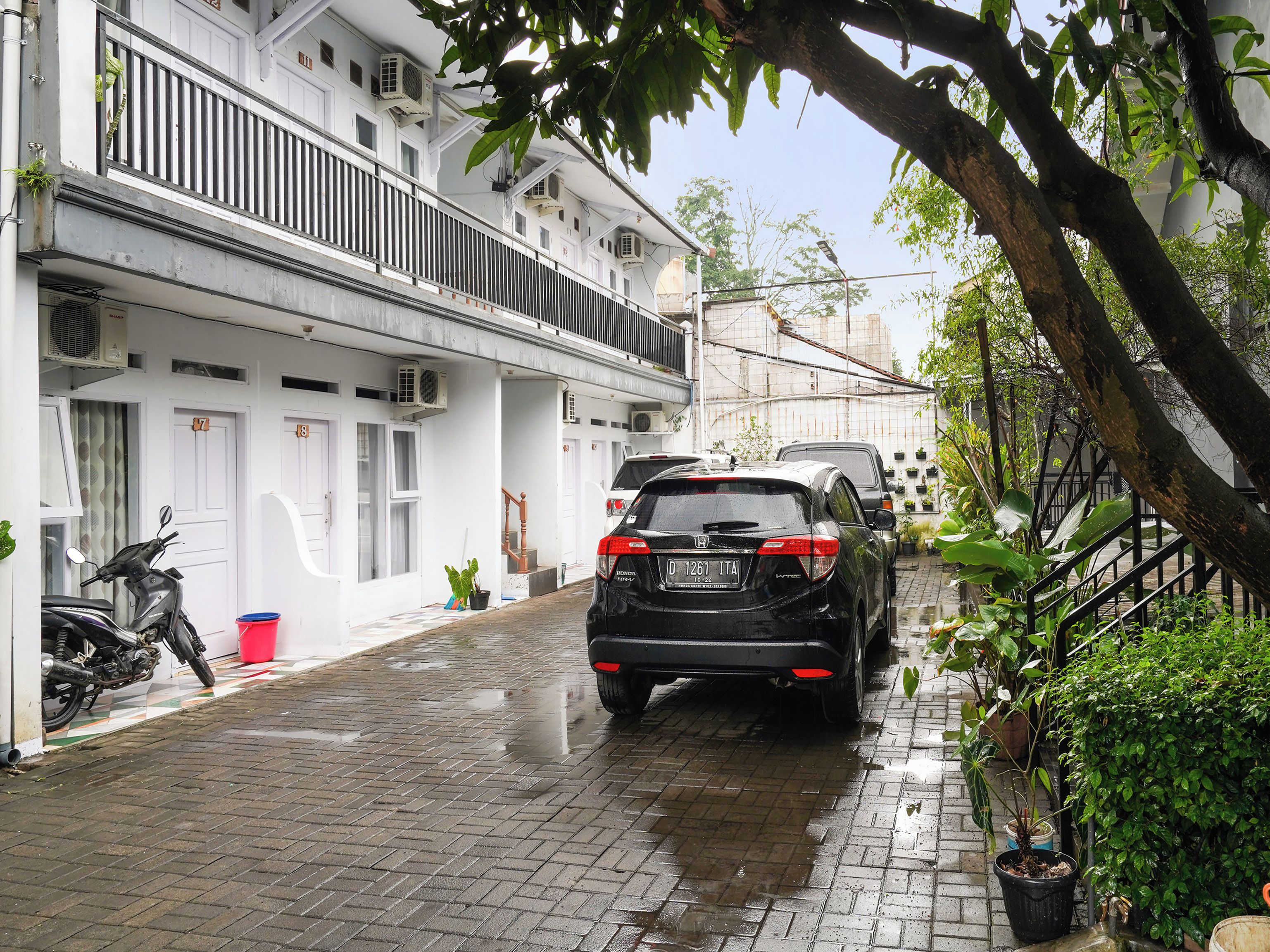 A narrow street with parked cars and a motorcycle, white buildings with balconies, and potted plants.