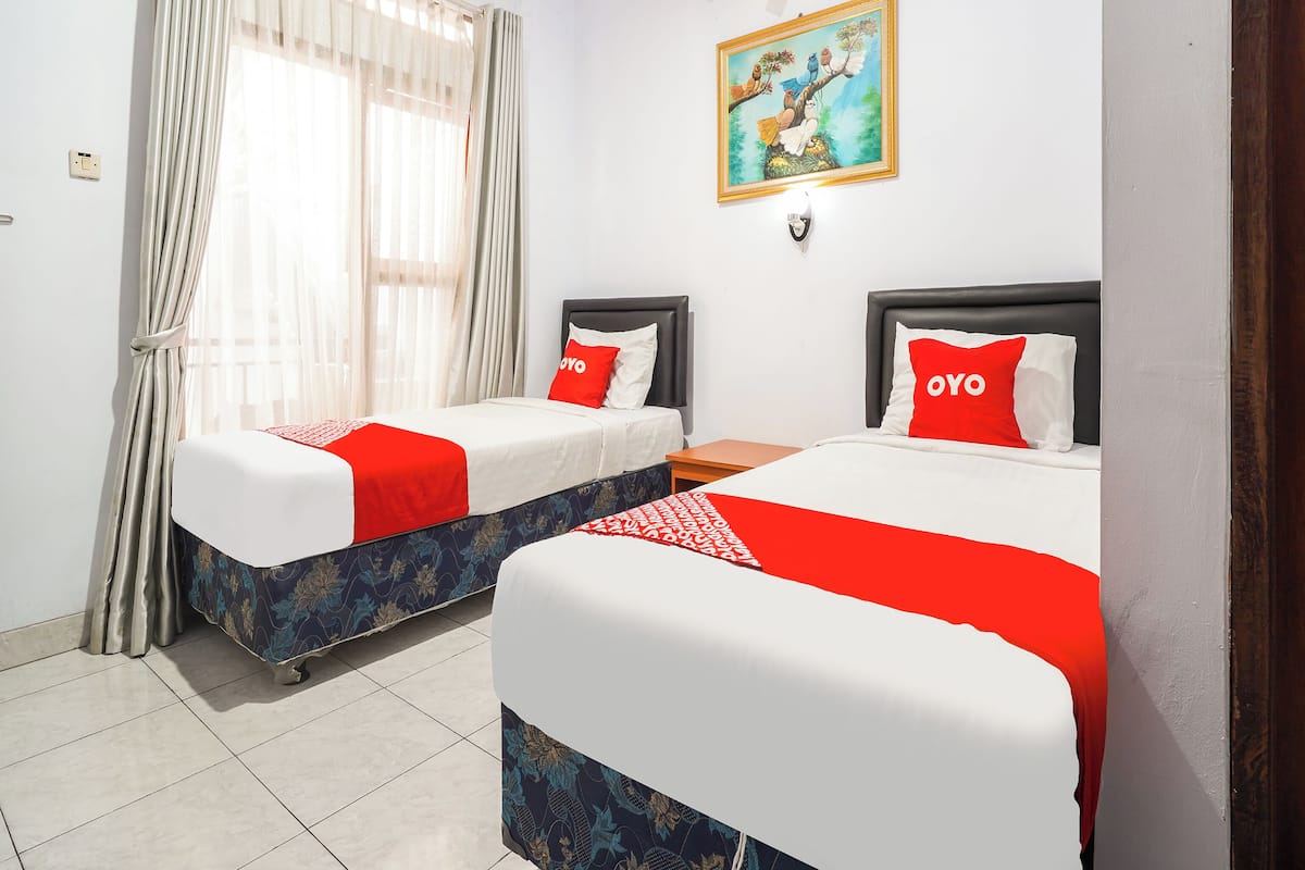 A hotel room with two beds, each with a red and white bedspread, a painting on the wall, and an air conditioning unit.
