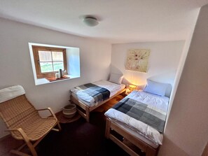 Una habitación pequeña y acogedora con dos camas individuales, una ventana con vistas, una silla y una lámpara de pared.
