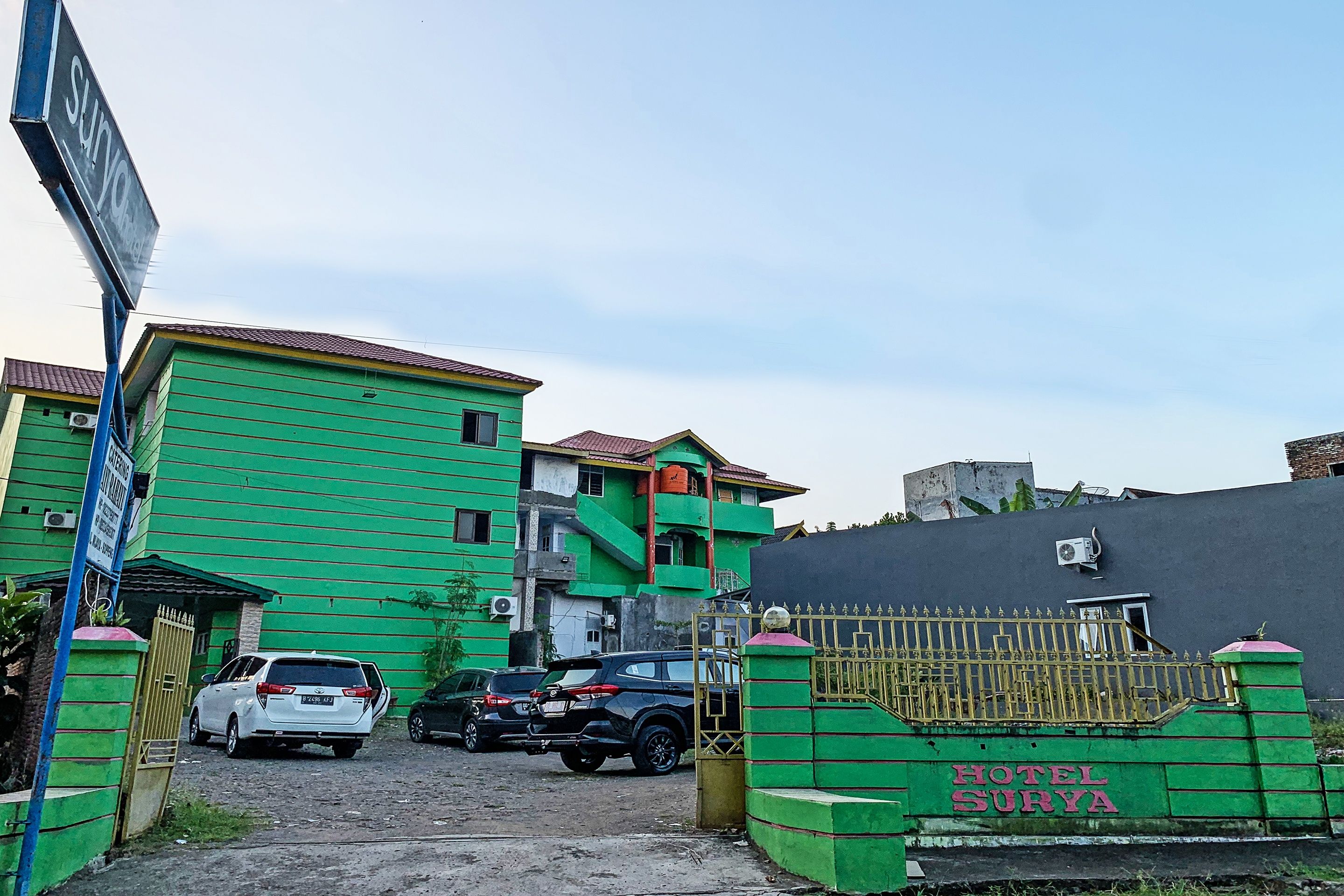 A hotel entrance with a gated driveway, a green building, and a sign reading 'HOTEL SURYA'.