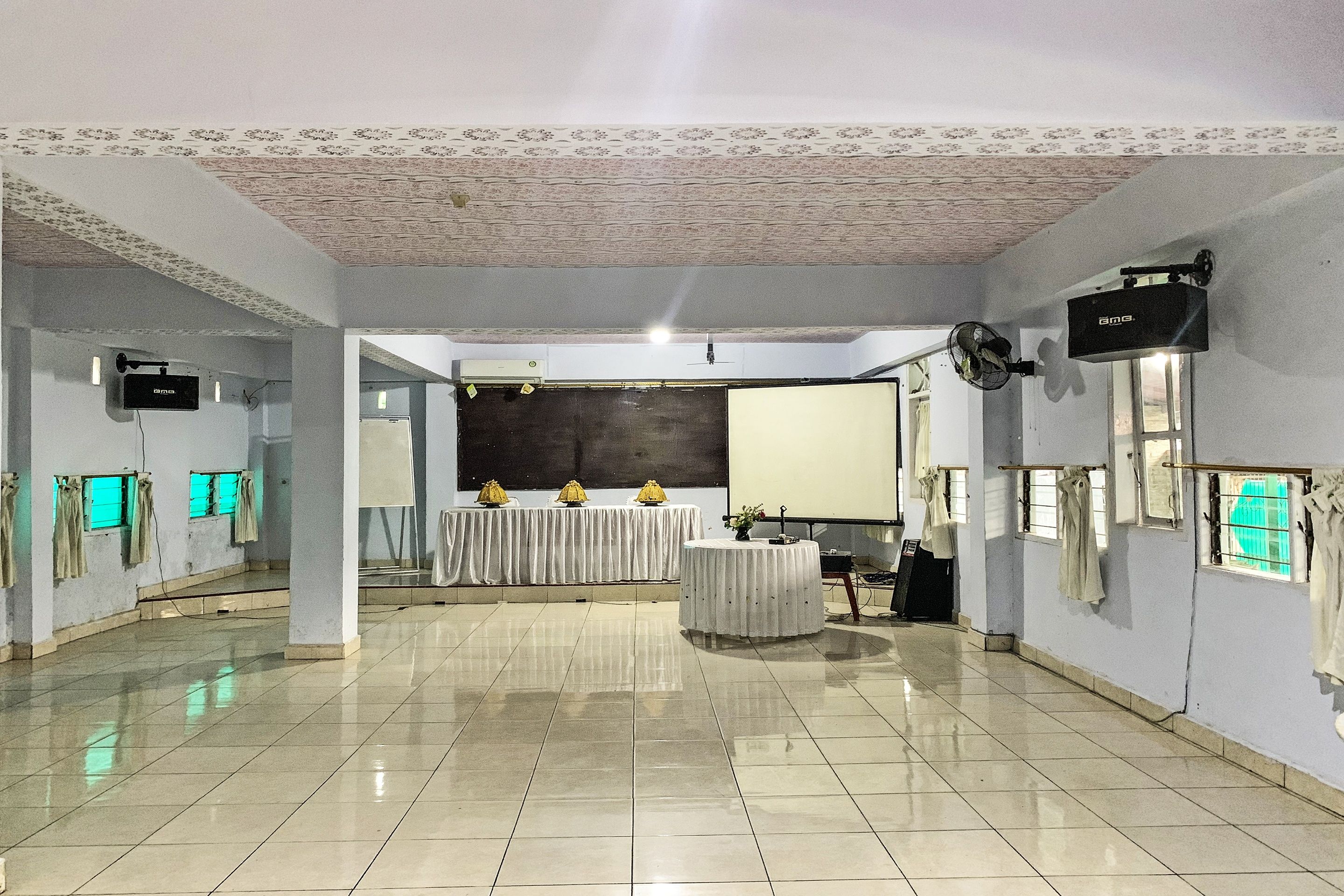 A spacious room with a tiled floor, a projector screen, a ceiling fan, and a table with a white tablecloth.