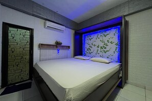 A bedroom with a bed, a window with a geometric pattern, an air conditioning unit, and a wall with a leaf pattern illuminated by blue light. - Oyo 91757 Surya Hotel (Watansoppeng)