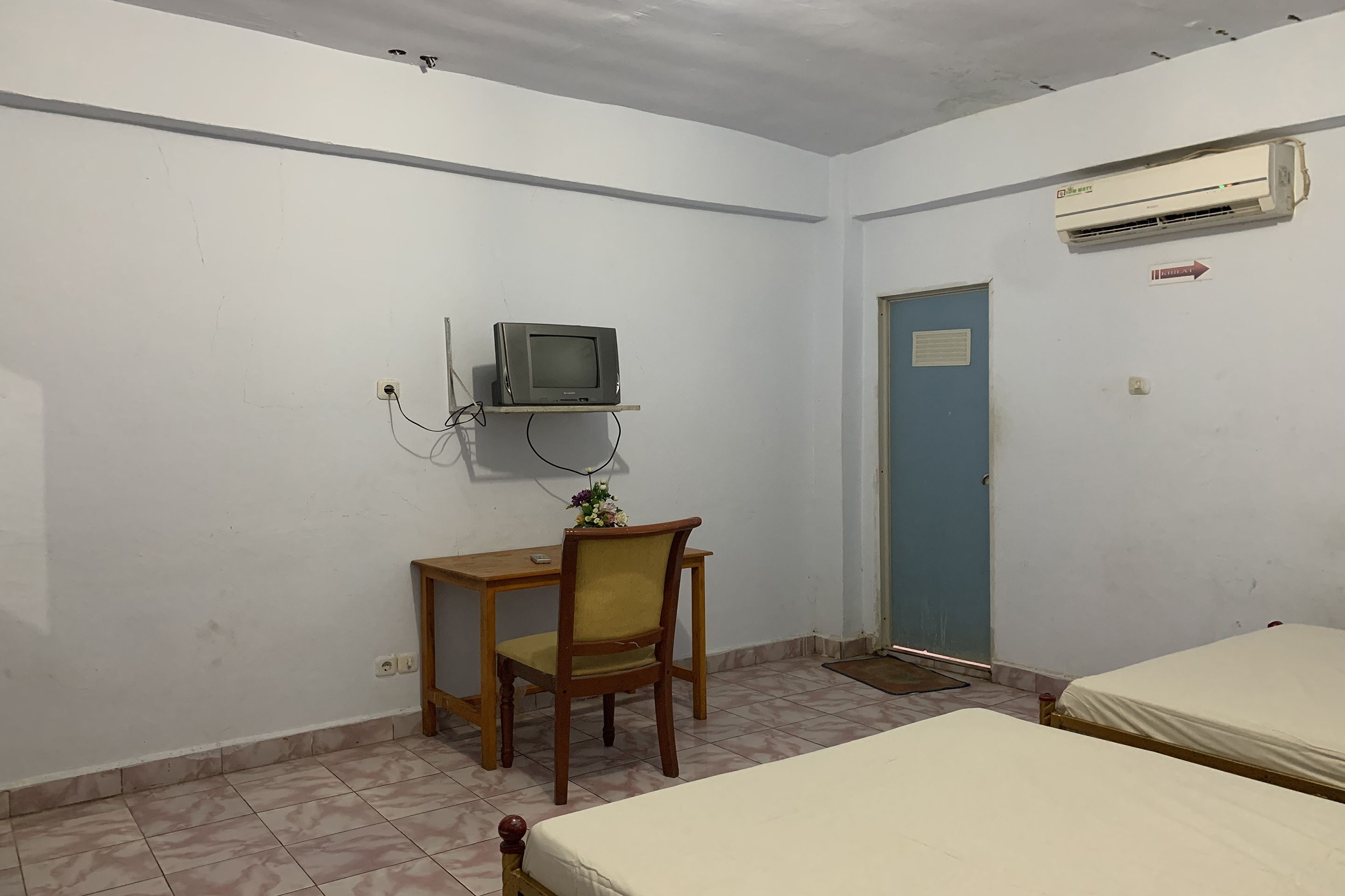 A room with two beds, a desk with a chair, a television mounted on the wall, and an air conditioning unit.