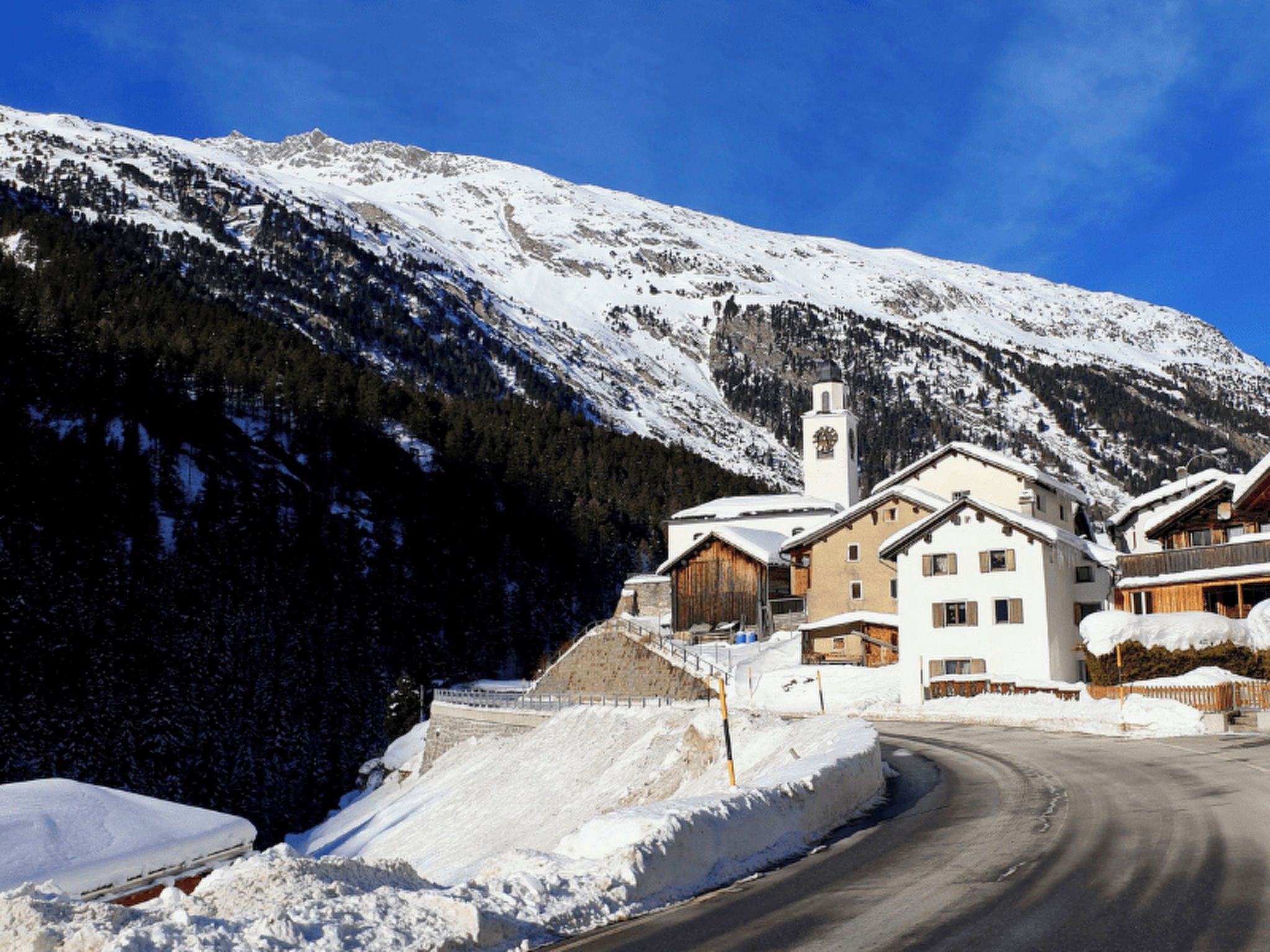 Un village de montagne enneigé, avec une église, des bâtiments en bois et une route sinueuse.