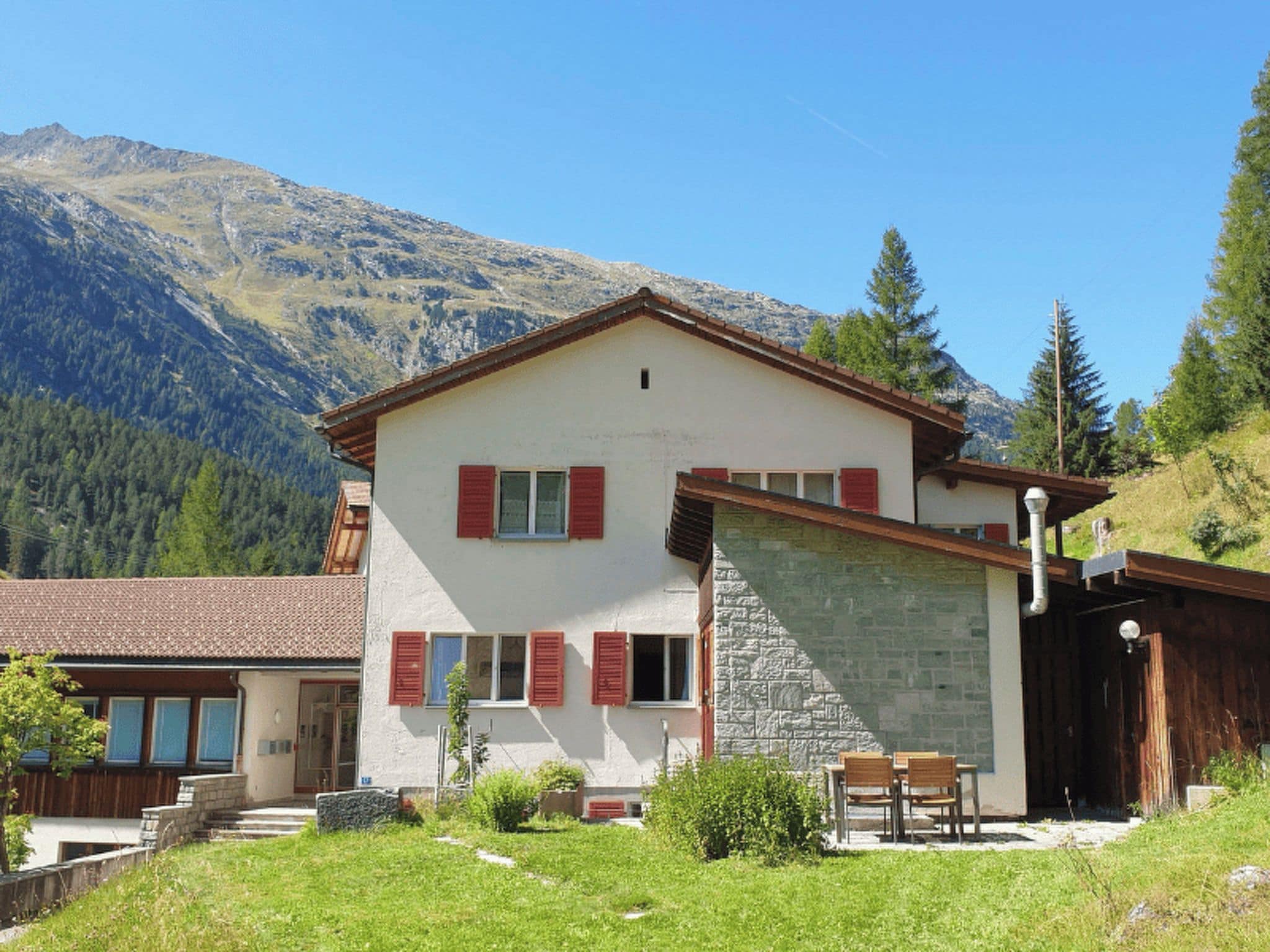 A two-story building with a stone facade and red shutters, set against a mountainous backdrop.