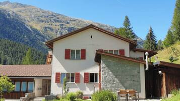 A two-story building with a stone facade and red shutters, set against a mountainous backdrop.