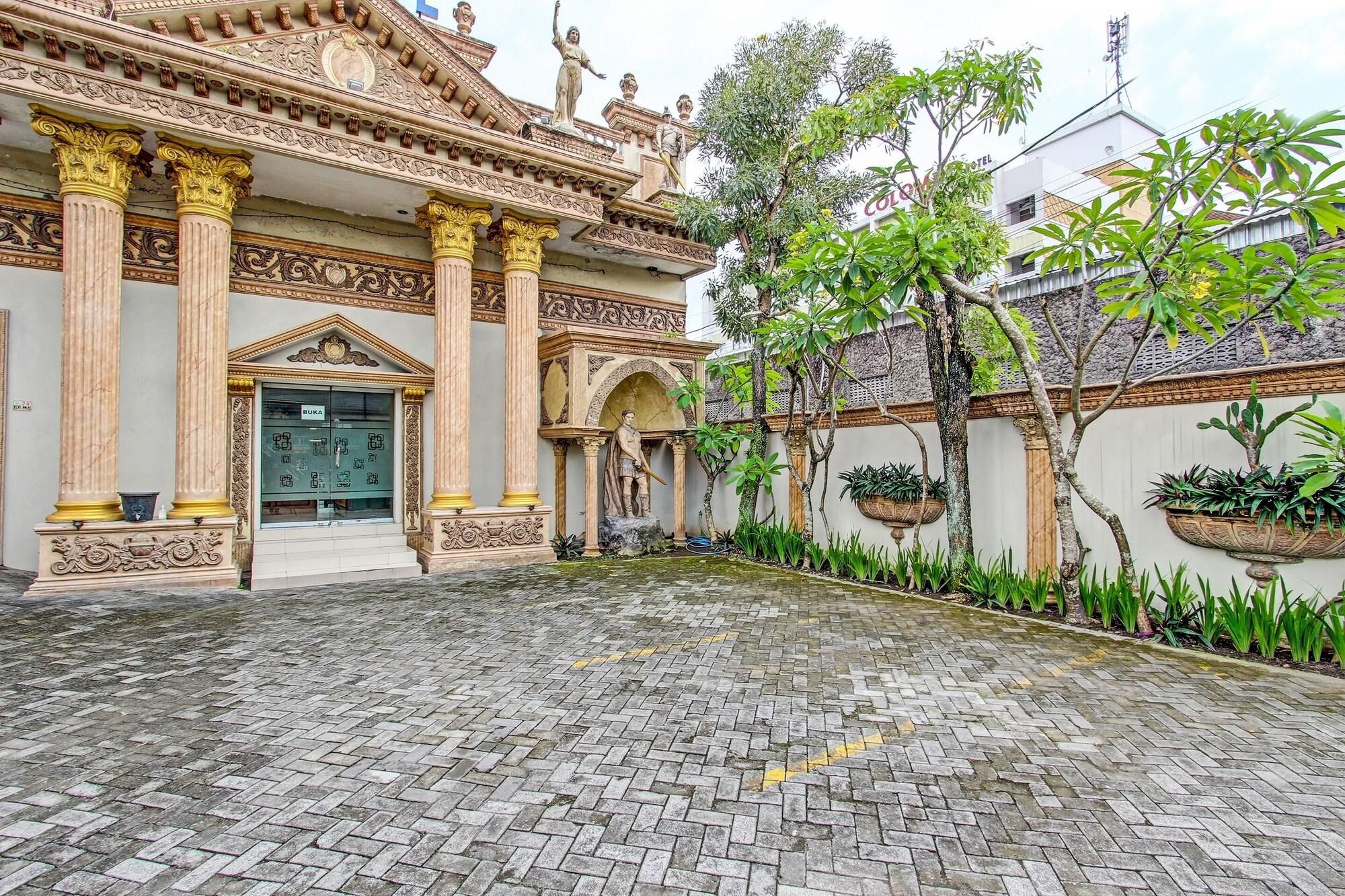 A courtyard with a traditional building featuring ornate columns and statues, a paved walkway, and potted plants.