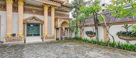 A courtyard with a traditional building featuring ornate columns and statues, a paved walkway, and potted plants.