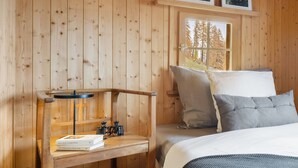 Ein Zimmer in einer Holzhütte mit einem Bett, einem Nachttisch mit Büchern und Fernglas sowie gerahmten Bildern an der Wand.