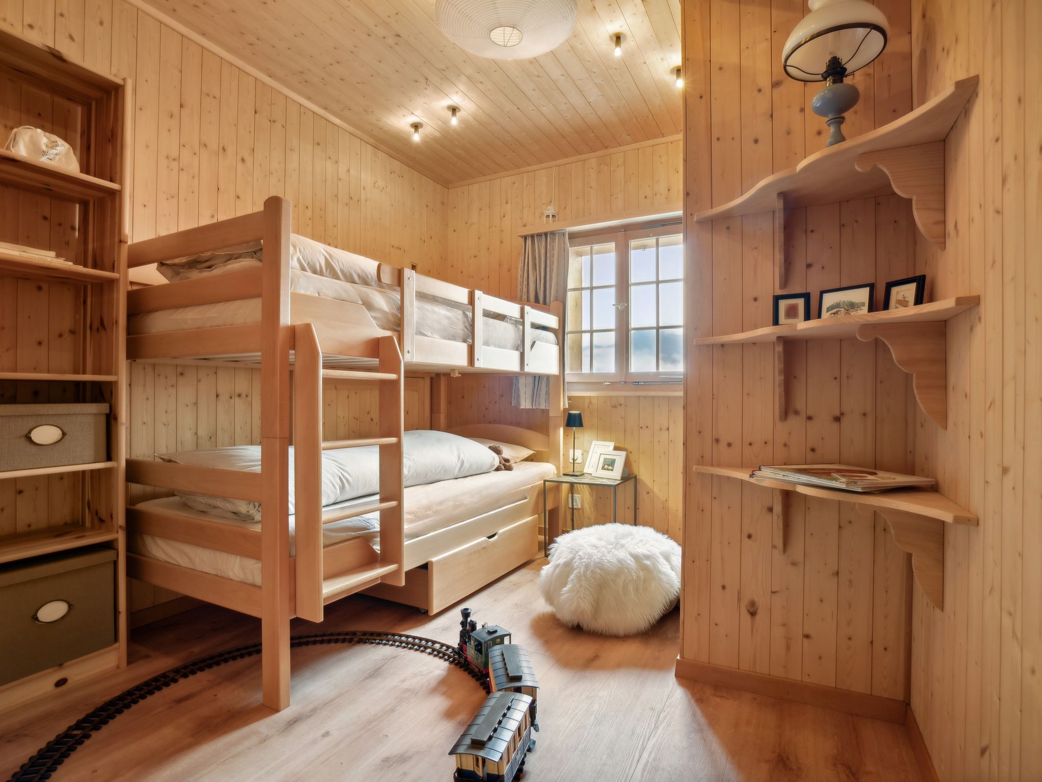 A wooden room with bunk beds, a small table with a lamp, a window, and a shelf with books.