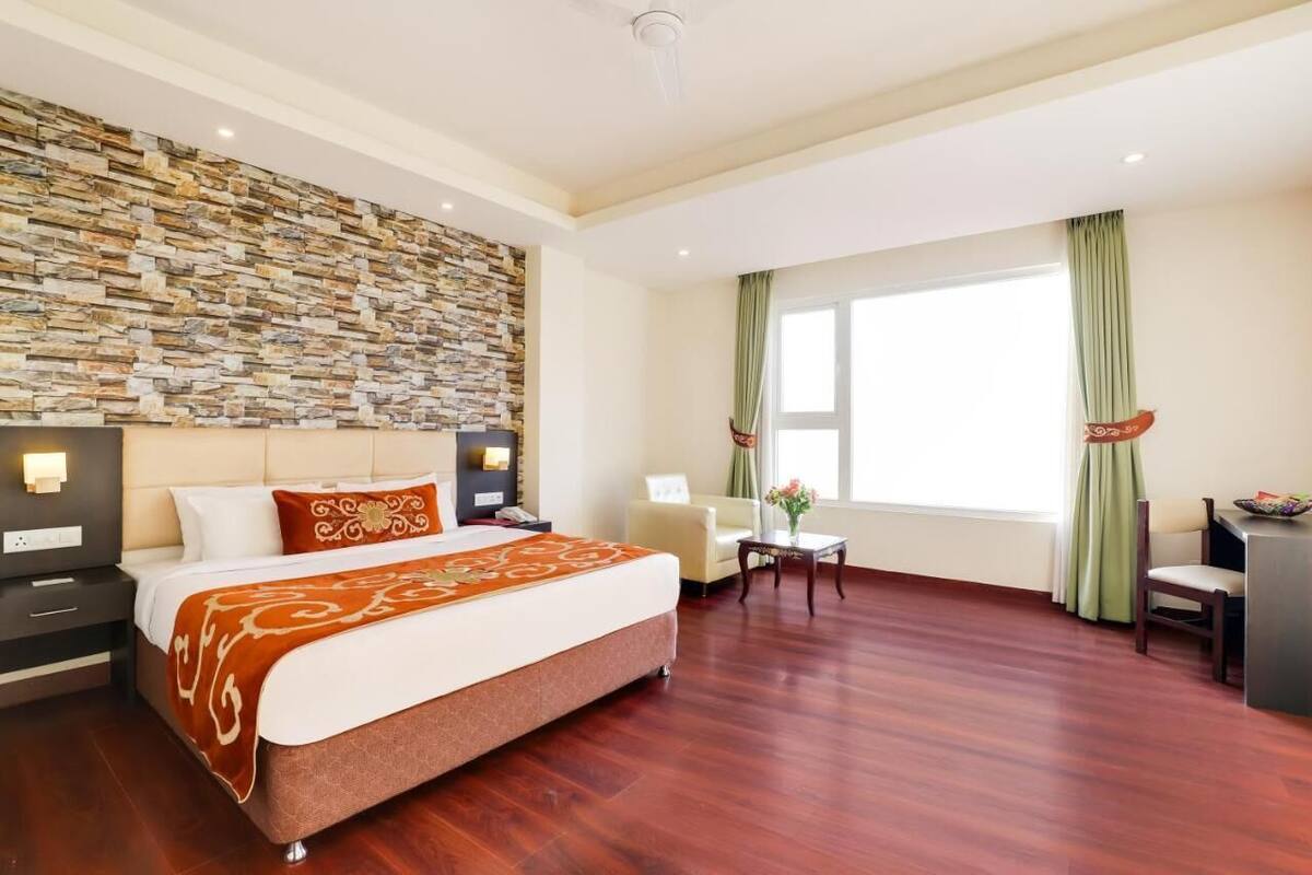 Una habitación de hotel con pared de piedra, una cama con colcha marrón, un escritorio con silla y una ventana con cortinas.