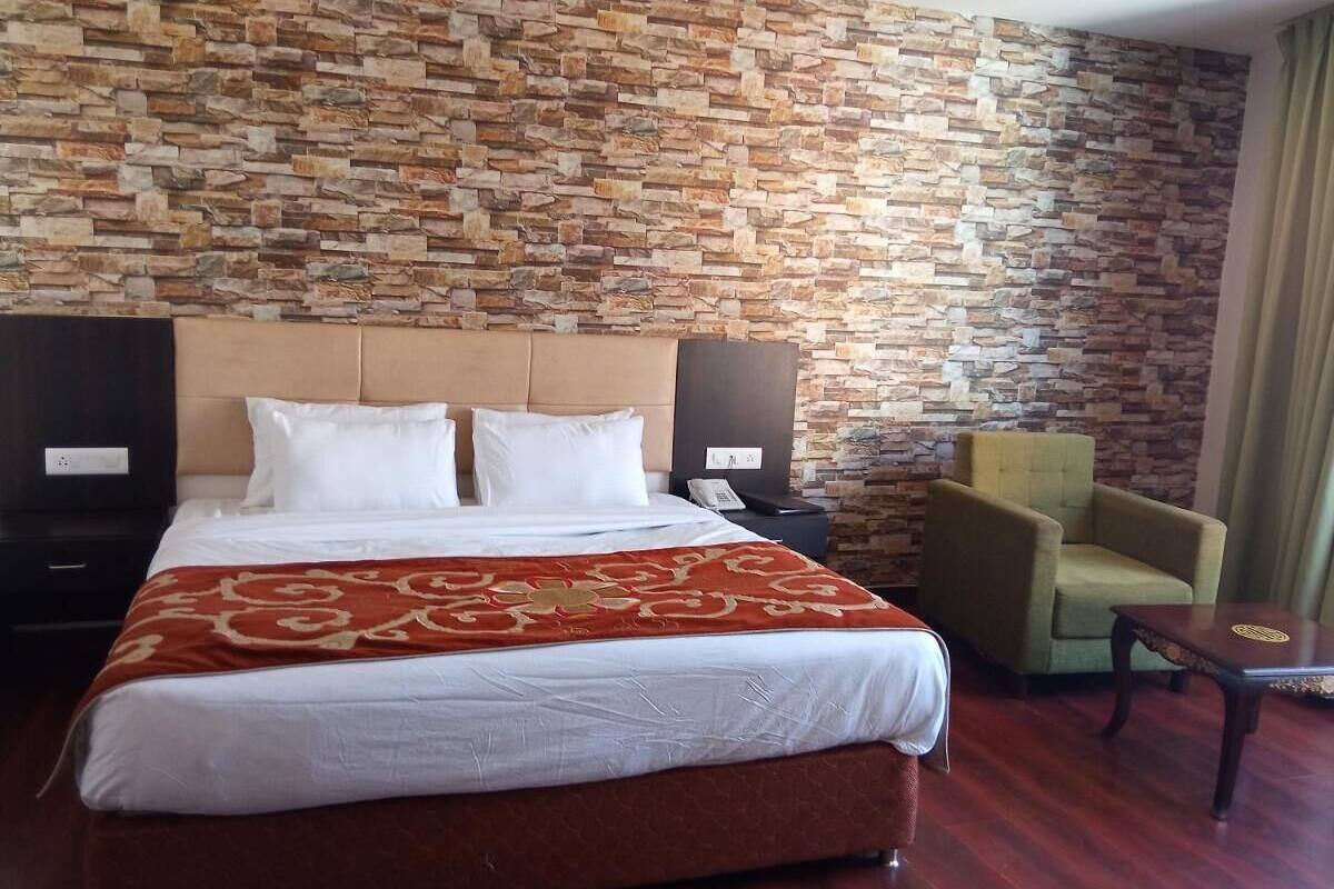 Una habitación de hotel con una cama grande, una pared de piedra, un sillón verde, una mesita pequeña y una mesita de noche con un teléfono.