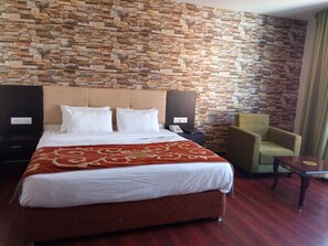 Une chambre d’hôtel avec un grand lit, un mur en pierre, un fauteuil vert, une petite table et une table de chevet avec un téléphone.