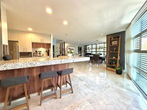 A modern kitchen with a marble countertop, wooden stools, and a dining area with a large window. - Penthouse with outdoor terrace in Lomas / Polanco (Ciudad de México)