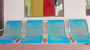 A row of blue metal chairs with perforated backs, positioned in front of a wall with a large mirror reflecting colorful curtains. - Oyo 91766 Fortuna Hotel Syariah (Banyumas)