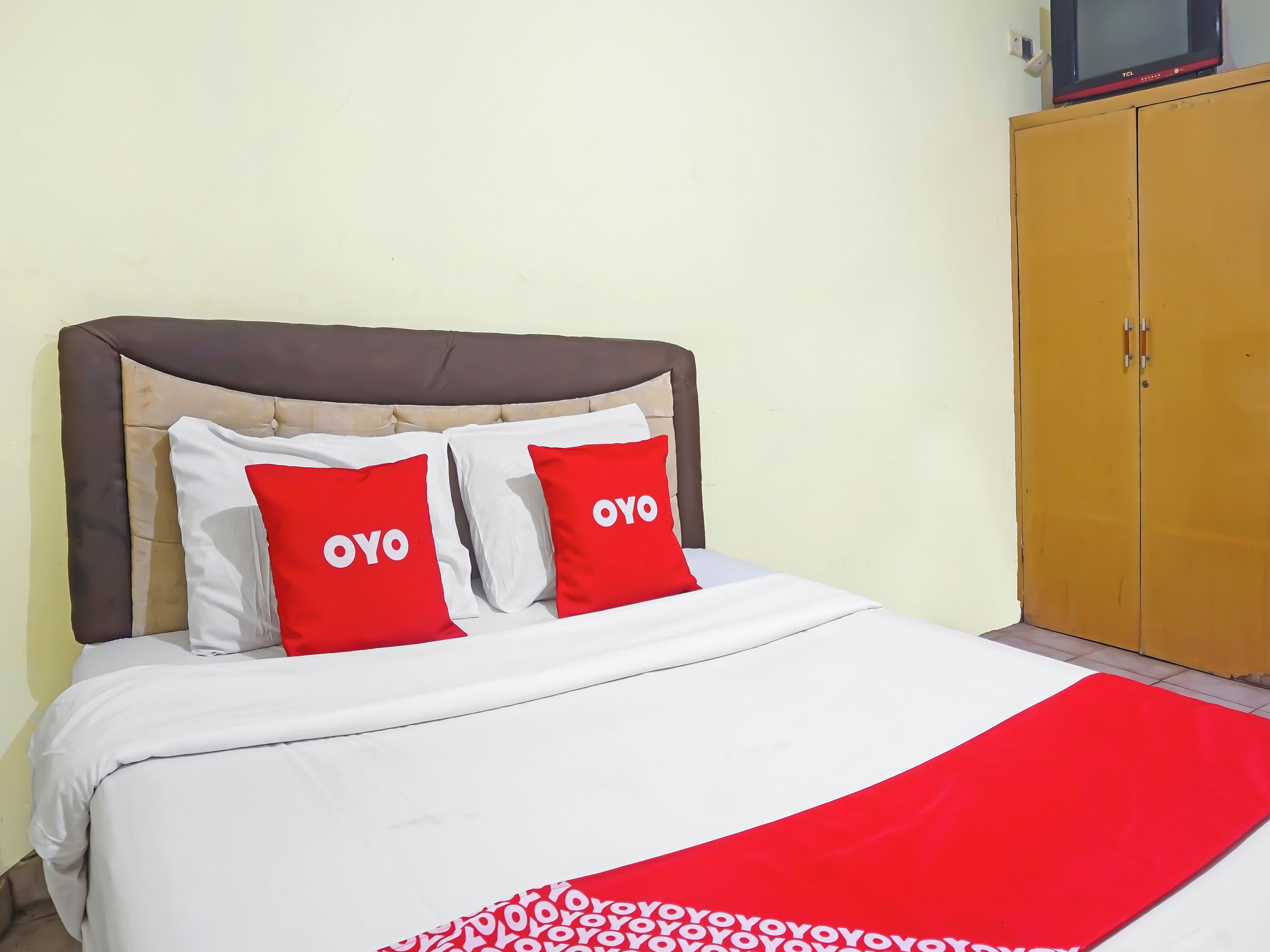 A bed with a white and red bedspread, two red pillows with 'OYO' text, a brown upholstered headboard, and a wooden wardrobe.