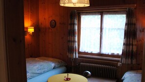 En stuga med träpanel, inredd med två sängar, ett bord, ett fönster med gardiner och en vägglampa.