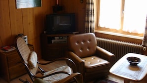 Ett rum med träpanel på väggarna, ett tv-appar, en bekväm fåtölj, ett litet bord med en skål och ett fönster med gardiner.