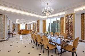 A spacious dining area with a long table set for a meal, surrounded by chairs, a chandelier, and large windows.