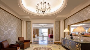 A spacious lobby with a chandelier, a seating area with leather chairs, a decorative console table with lamps, and a large mirror. - Dusit Hotel and Suites Doha (Doha)