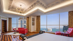 A hotel room with a large bed, a red sofa, a red armchair, a small table, and a view of the city. - Dusit Hotel and Suites Doha (Doha)
