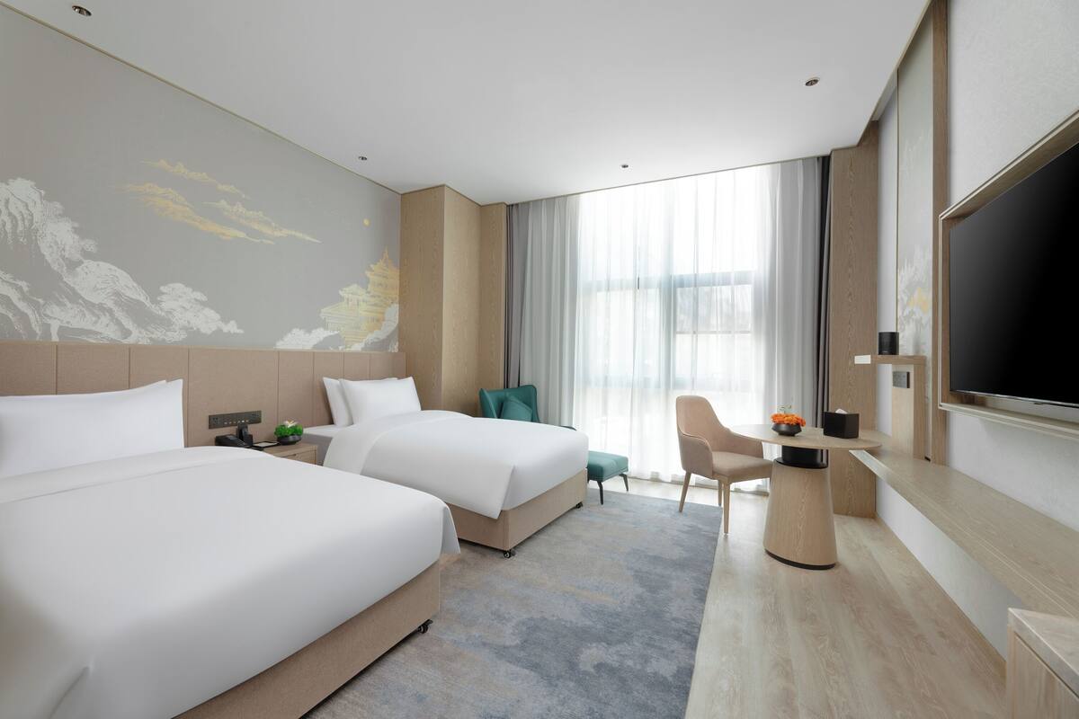 Ein Hotelzimmer mit zwei Betten, einem Schreibtisch, einem Fernseher und einem großen Fenster mit Vorhängen.