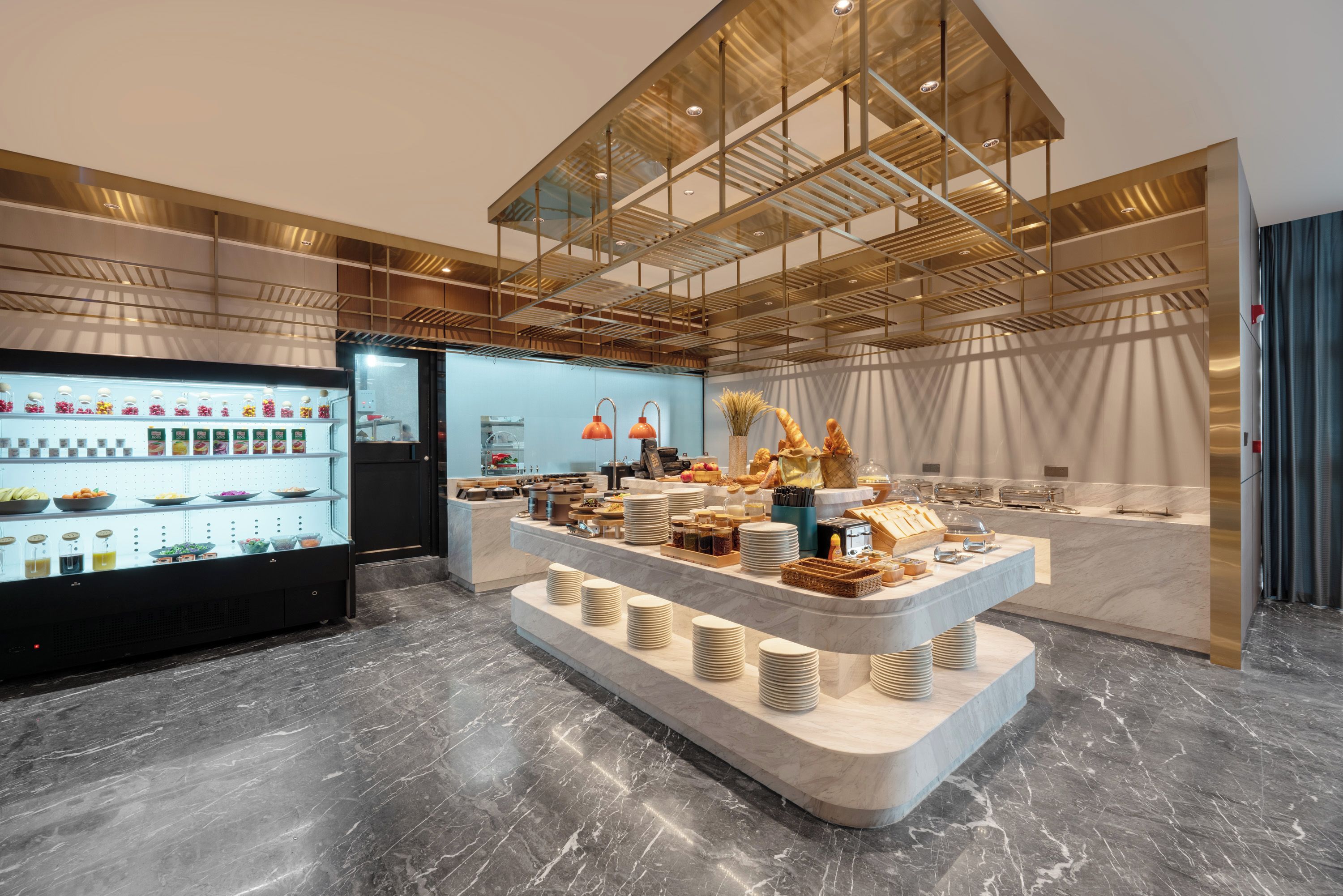 A modern hotel lobby with a marble floor, a glass display case with food items, and a central counter with plates and bowls.