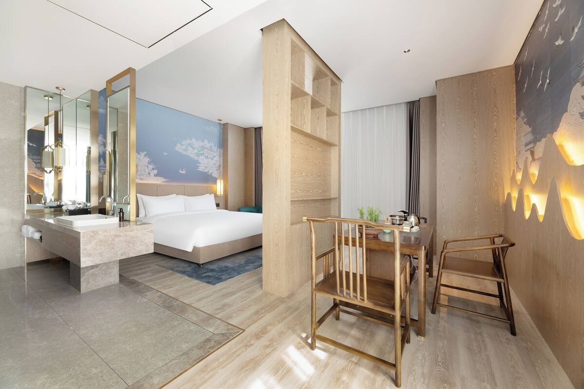 Ein modernes Hotelzimmer mit einem großen Bett, einer Sitzecke mit Tisch und Stühlen, einem Badezimmer mit Waschbecken und Spiegel sowie einem großen Wandbild.