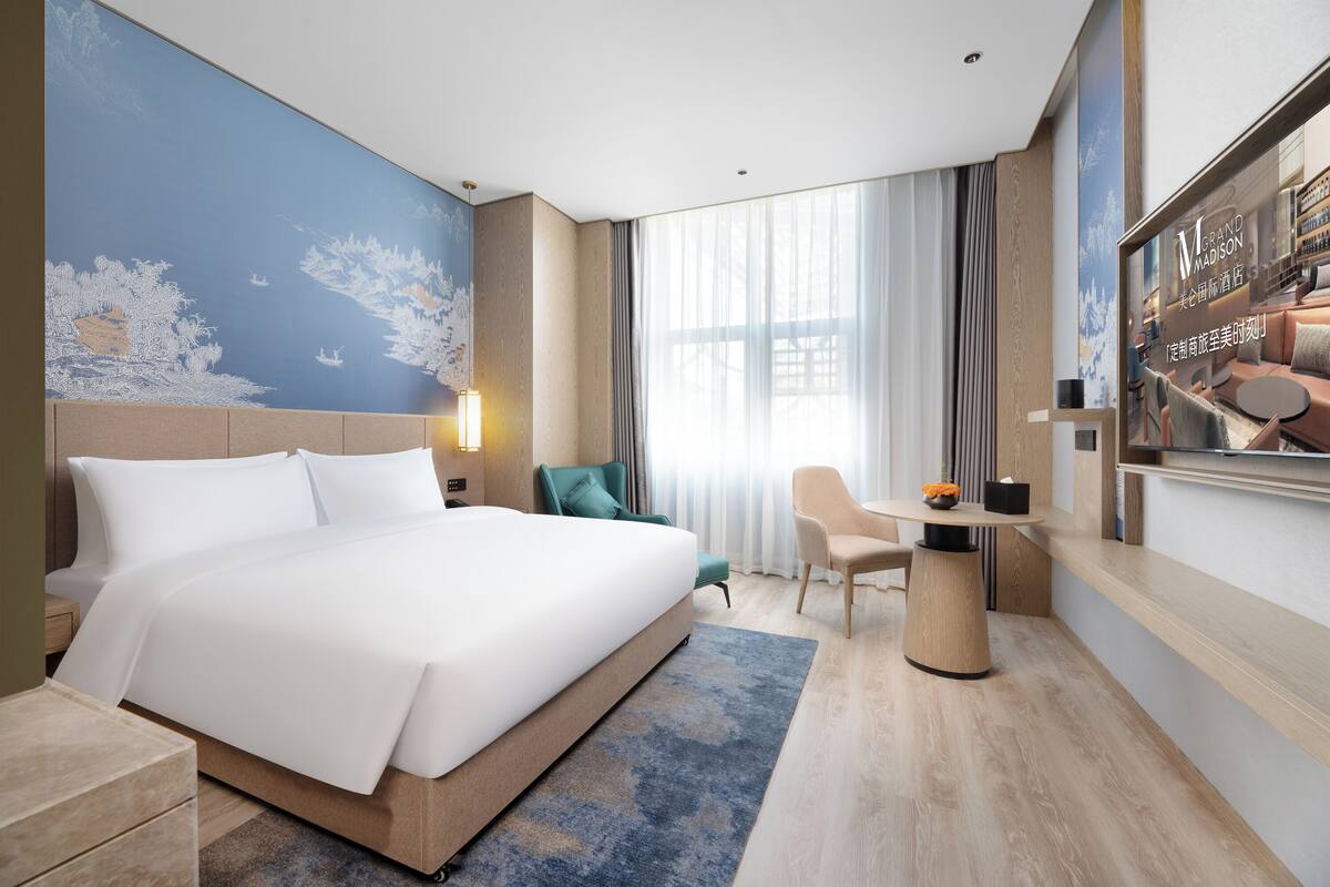 Ein Hotelzimmer mit einem großen Bett, einem Schreibtisch, einem Sessel, einem Fernseher und einem Wandgemälde.