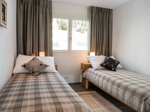 Une chambre avec deux lits, chacun recouvert d’une couverture à carreaux et agrémenté d’oreillers assortis, une fenêtre avec des rideaux et un petit tabouret en bois.