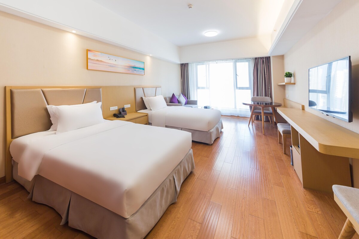 Habitación de hotel con dos camas, un escritorio, un televisor y un ventanal grande.