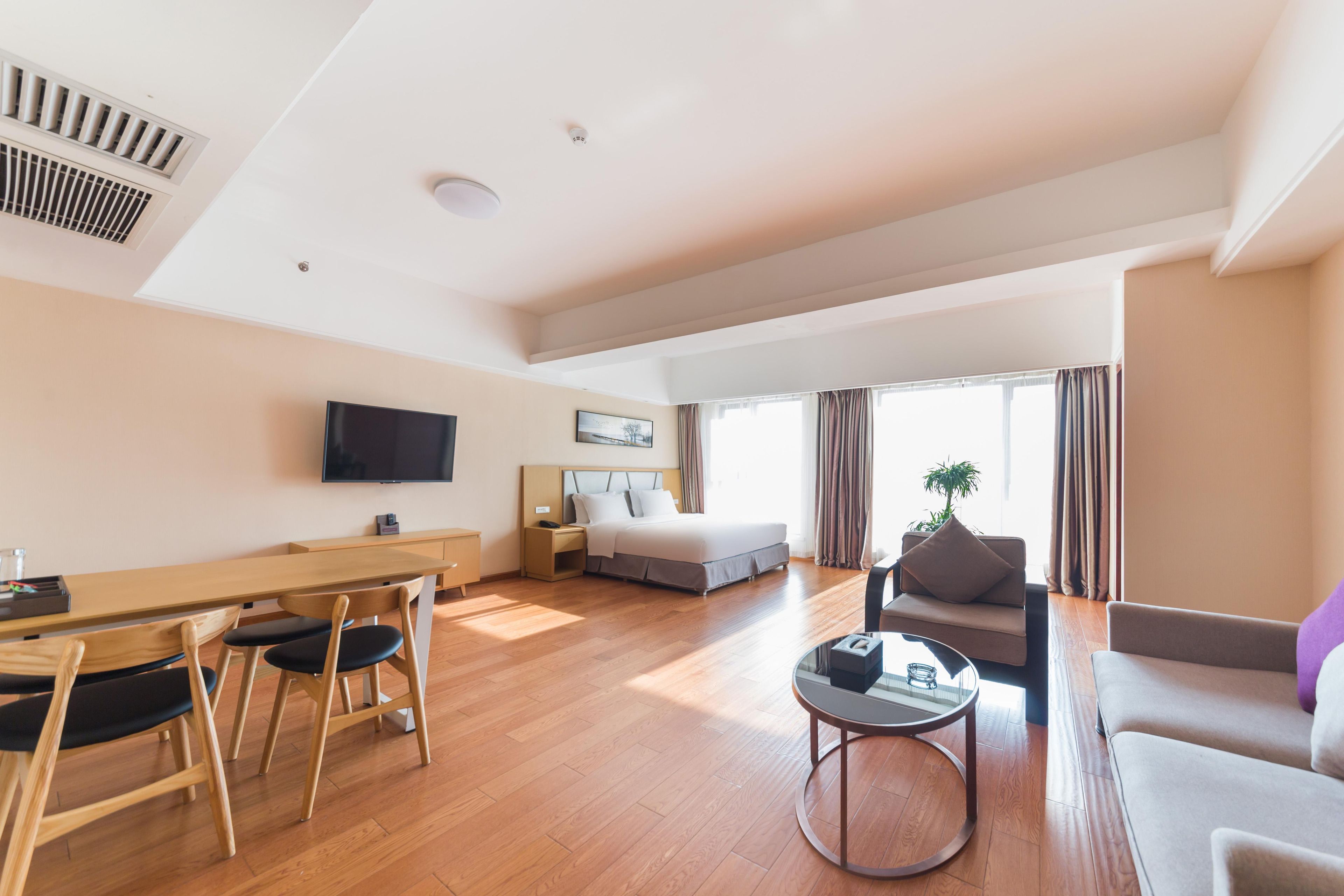 Una habitación de hotel moderna con una mesa de comedor de madera, un sofá, una cama, un televisor y un ventanal con cortinas.