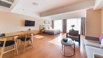 Una habitación de hotel moderna con una mesa de comedor de madera, un sofá, una cama, un televisor y un ventanal con cortinas.