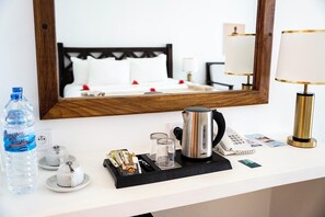 Habitación de hotel con cama, mesita de noche con tetera, lámpara, botella de agua y bandeja con artículos de aseo.