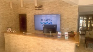 A reception desk with a flat-screen TV displaying 'ALLADIN' branding, a ceiling fan, and a brick wall backdrop. - Alladin Boutique Beach Hotel And Spa Zanzibar (Matemwe)