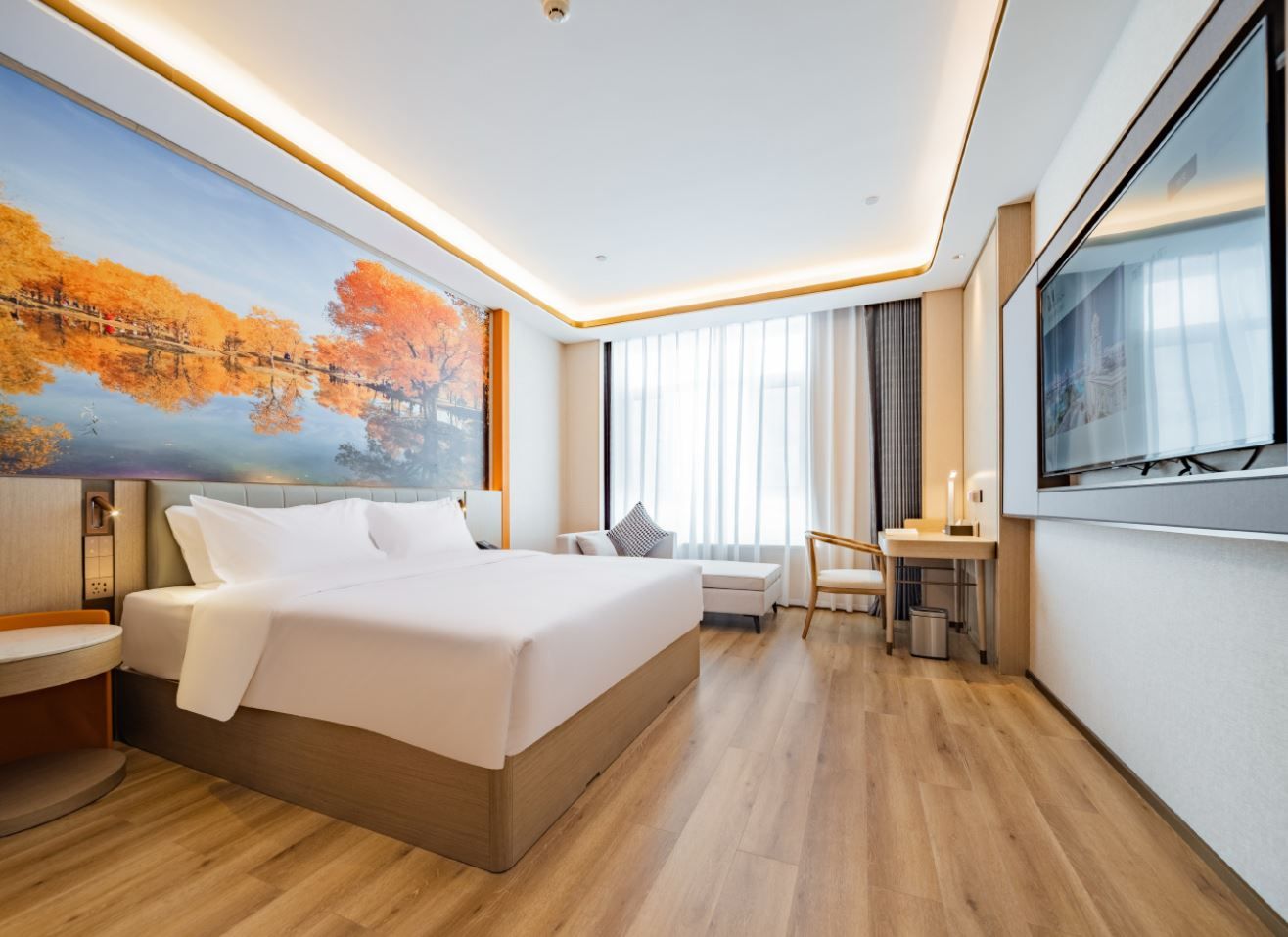 A modern hotel room with a large bed, a desk, a chair, a TV, and a painting of autumn trees.
