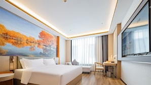 A modern hotel room with a large bed, a desk, a chair, a TV, and a painting of autumn trees.