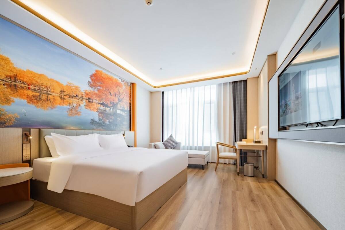 Una habitación de hotel moderna con una cama grande, un escritorio, una silla, un televisor y un cuadro de árboles otoñales.