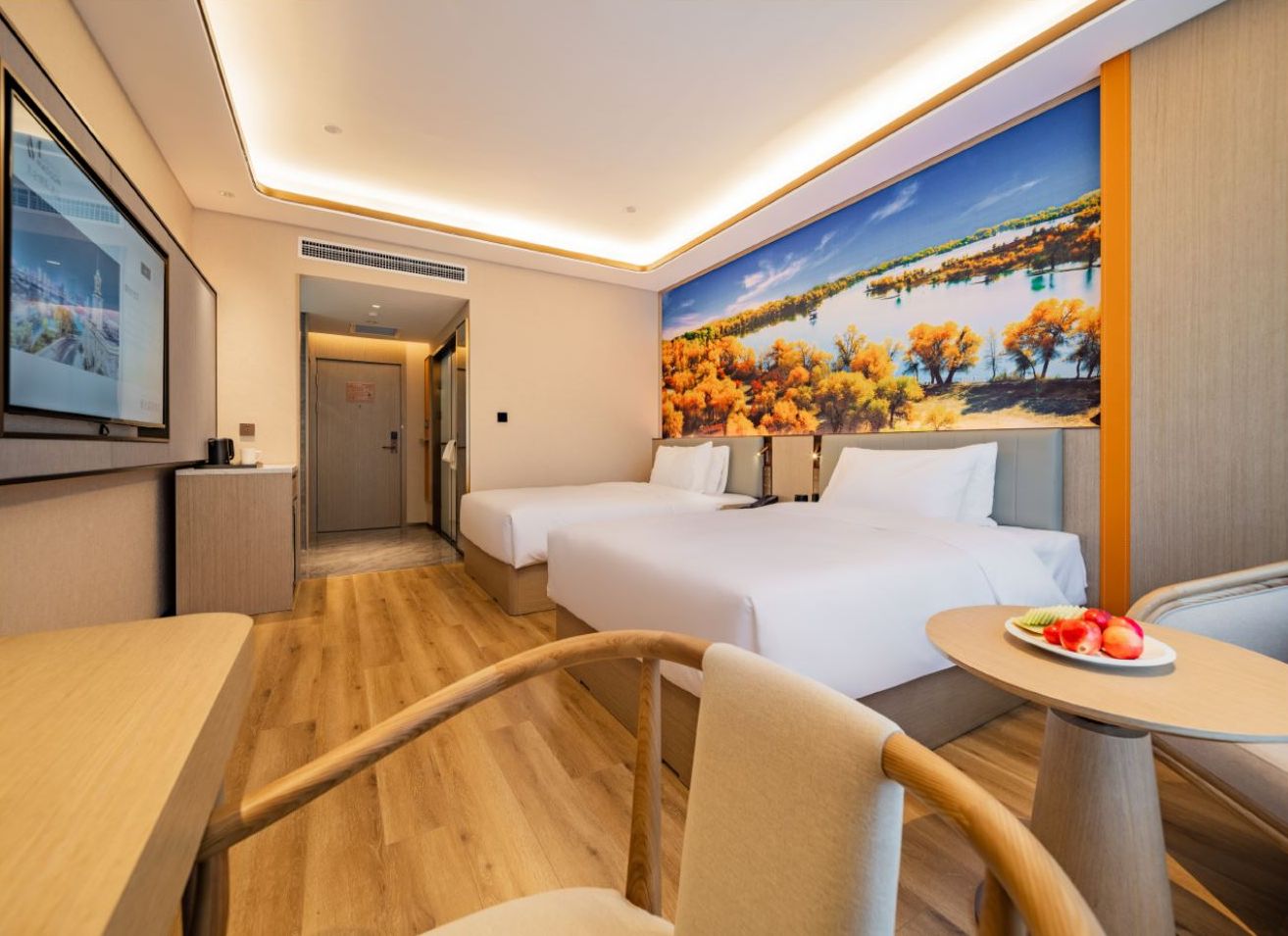 A hotel room with two beds, a dining table, a chair, a small table with fruit, and a mural of autumn trees.
