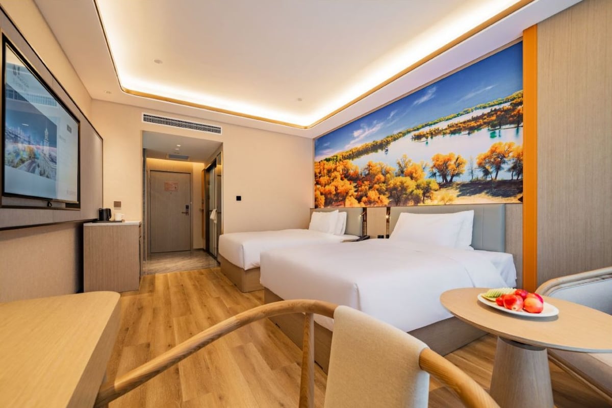 Habitación de hotel con dos camas, una mesa de comedor, una silla, una mesita con fruta y un mural de árboles otoñales.